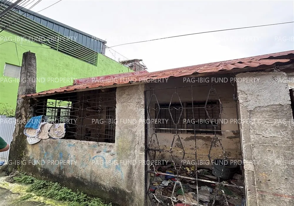 Front of the single-storey row house in Belvedere Towne I, Tanza, Cavite, with a rusty tiled roof, concrete walls with graffiti and a metal-grille front gate, photographed for the Pag-IBIG auction listing